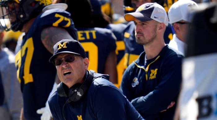 Michigan head football coach Jim Harbaugh looks on from the sideline in confusion while analyst Connor Stalions watches from behind him.
